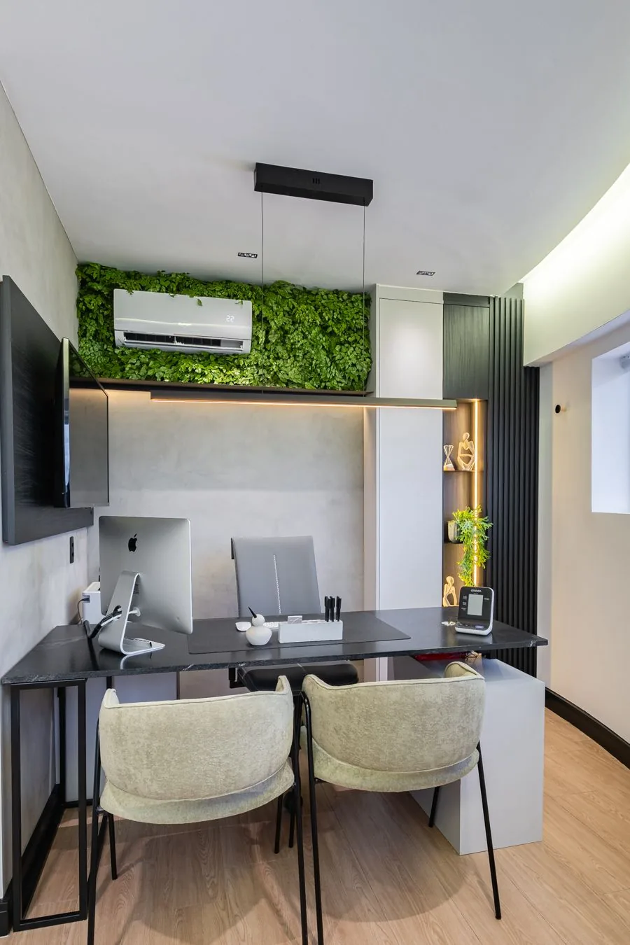 Consultório visto da entrada, com mesa preta, computador e poltronas bege sob a parede de folhagem verde