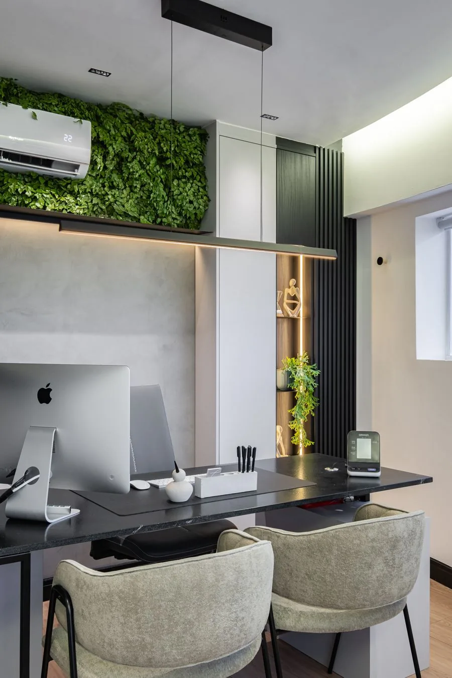 Consultório com mesa preta, dois iMacs e poltronas bege sob a parede de folhagem verde