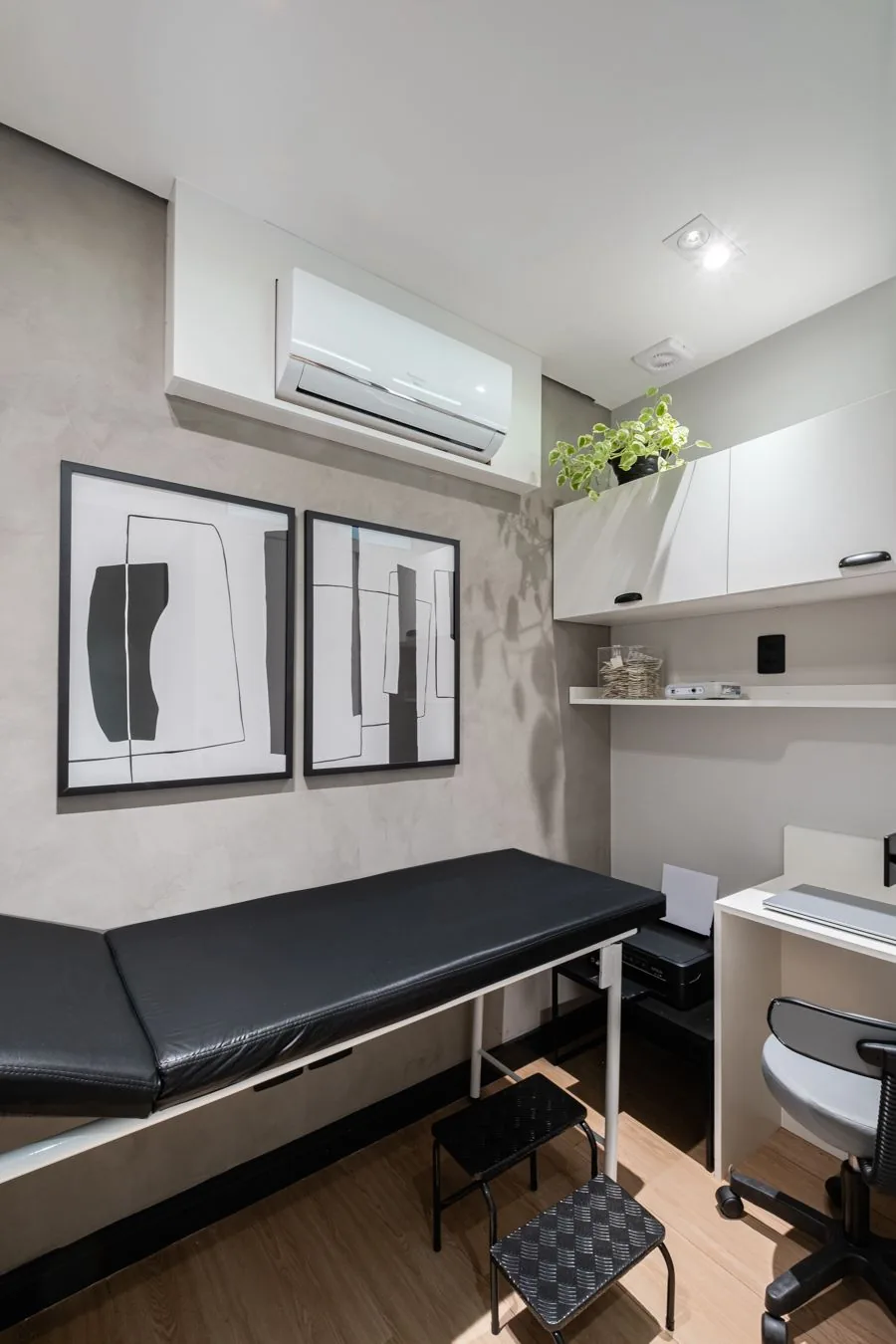 Sala de exame com maca preta, dois quadros na parede e armário branco